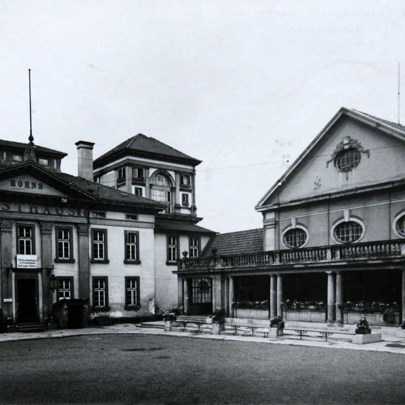 Ein altes Foto zeigt zwei historische Gebäude mit der Aufschrift "Rohns" und einer Terrasse mit Säulen.