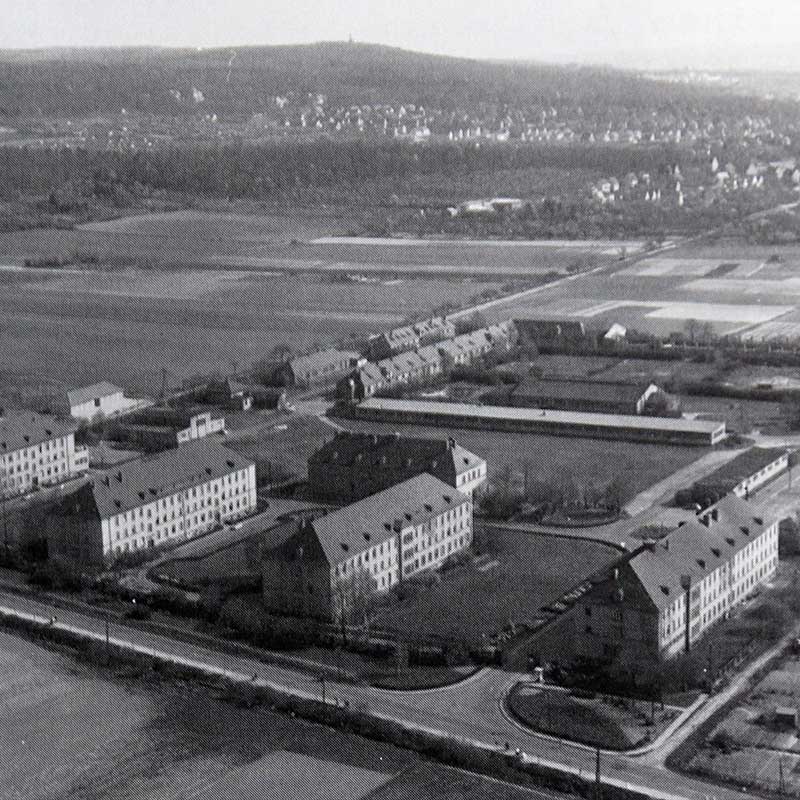 Luftaufnahme einer ländlichen Gegend mit Wohngebäuden, Straßen und Feldern im Hintergrund. Schwarz-Weiß-Bild.
