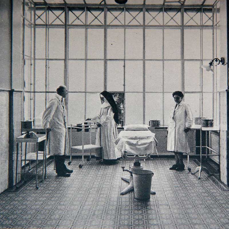 Ein Krankenhauszimmer mit drei Personen in weißen Kitteln, einem Bett und medizinischen Utensilien.