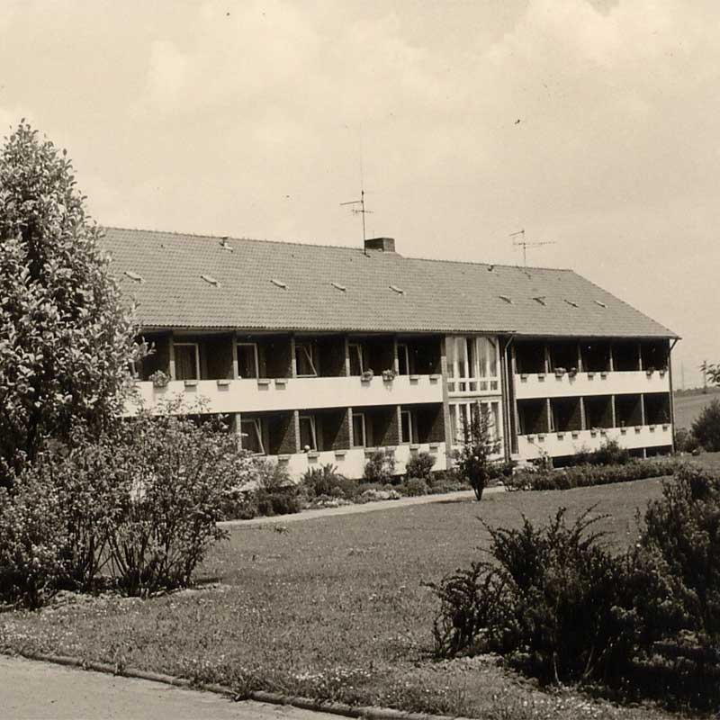 Ein zweistöckiges Gebäude mit mehreren Fenstern, umgeben von Bäumen und einem bewachsenen Vorgarten.