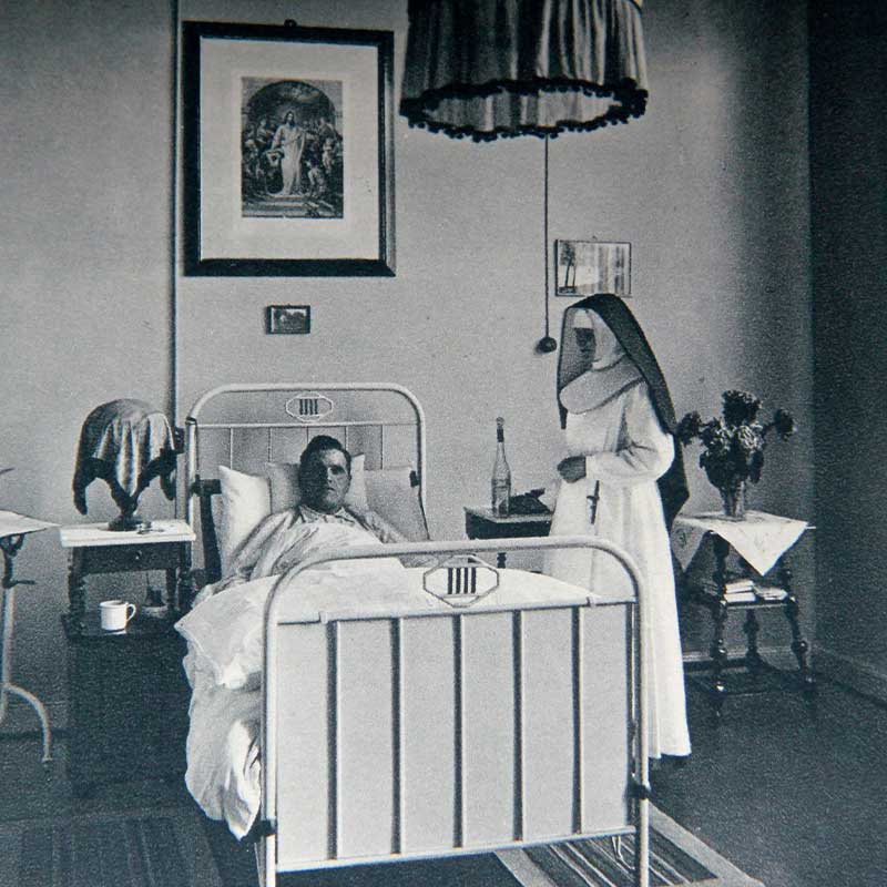 Ein Patient liegt im Bett, während eine Nonne neben ihm steht. Der Raum ist einfach eingerichtet.