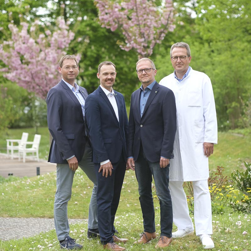 Vier Männer in Anzügen und ein Arzt im Garten mit blühenden Bäumen im Hintergrund.