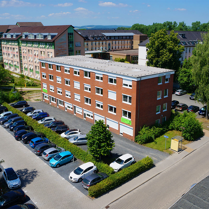 Rotes Backsteingebäude mit vielen Fenstern, umgeben von geparkten Autos und grüner Landschaft.