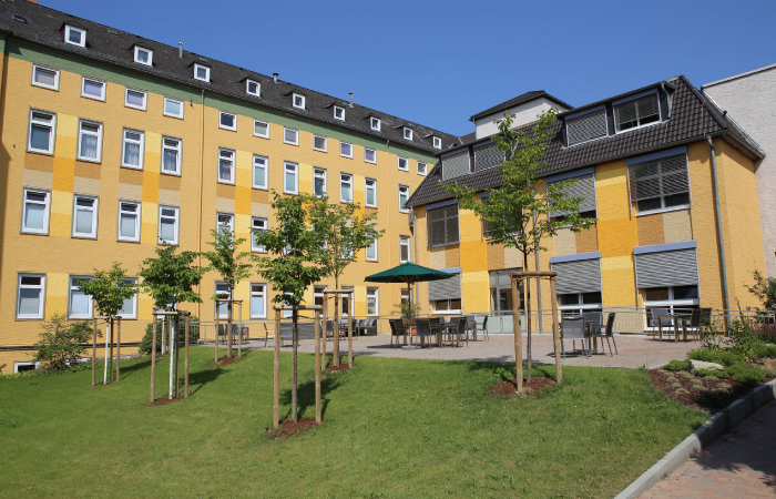 Ein gelbes Gebäude mit mehreren Fenstern und einem begrünten Innenhof mit jungen Bäumen und Sitzbereich.