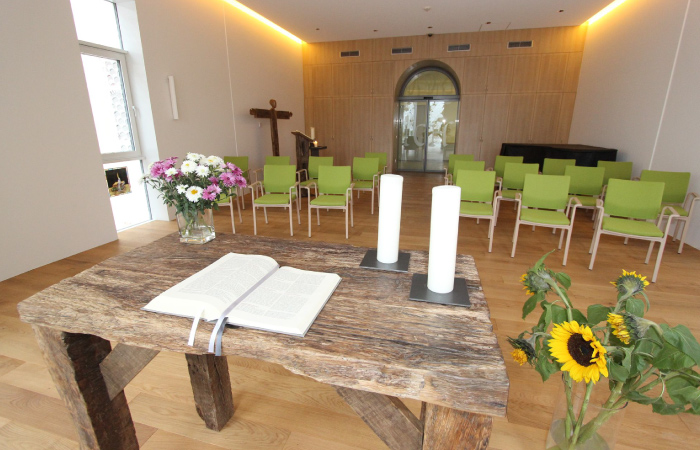 Ein Raum mit Tisch, Bibel, Kerzen, Blumen und grünen Stühlen, ein Kreuz im Hintergrund.