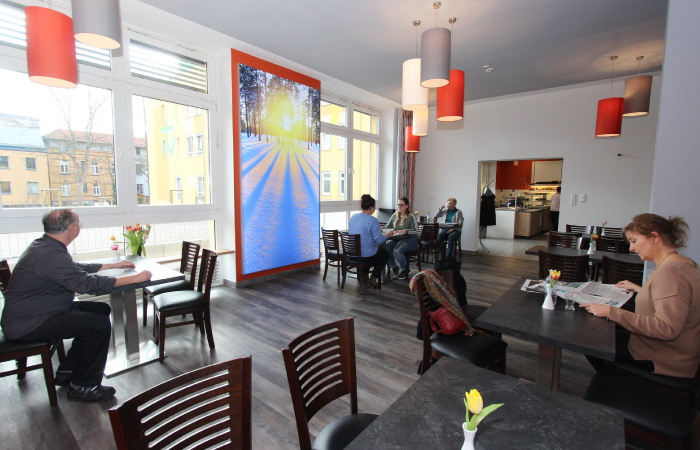 Ein Café mit Gästen, gemütlicher Atmosphäre, Tischen, Stühlen, großen Fenstern und einem großen Wandbild.