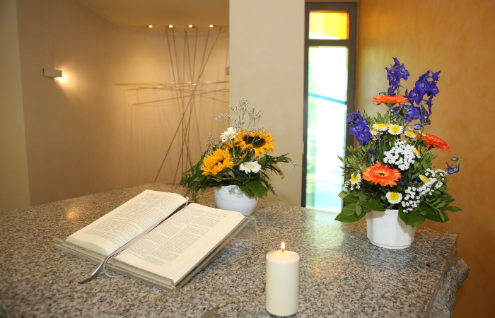 Ein aufgeschlagenes Buch, zwei Blumengestecke und eine brennende Kerze auf einem Tisch in einem hellen Raum.