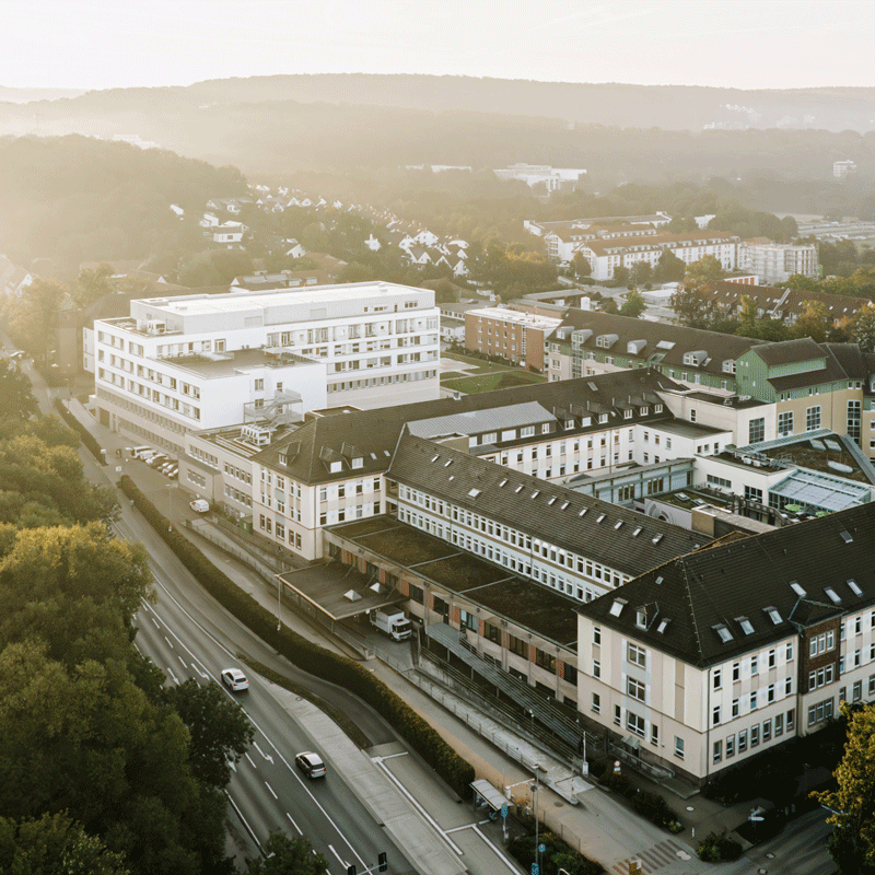 Luftaufnahme eines Krankenhauskomplexes in einer hügeligen, bewaldeten Landschaft.