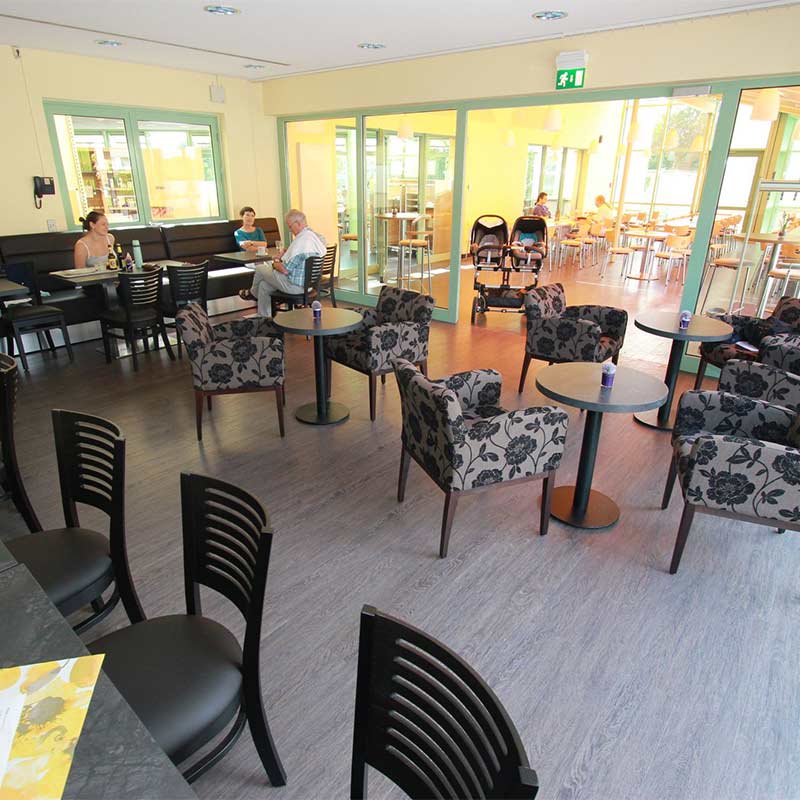 Ein Café mit Tischgruppen, Sesseln und Personen, einige leere Tische, warme Beleuchtung, ein Kinderwagen.