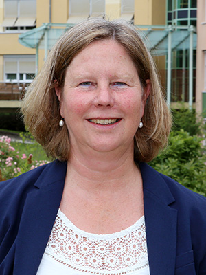 Es zeigt eine lächelnde Frau mit mittellangem Haar, weißer Bluse und Blazer vor einem Gebäude und Pflanzen.