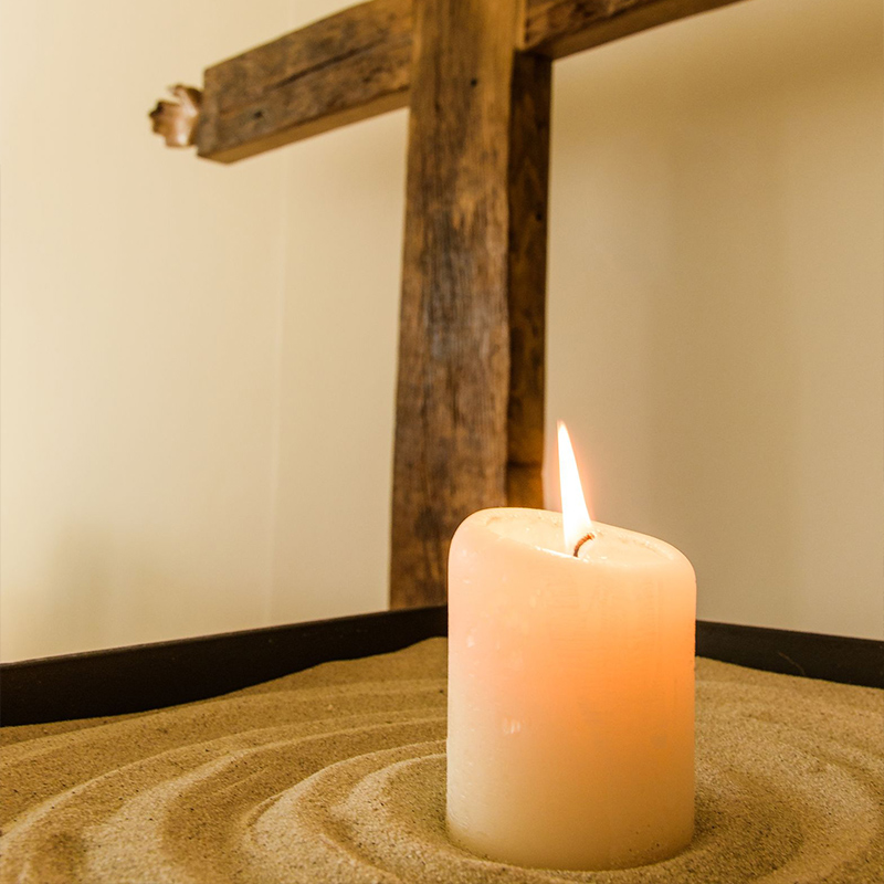 Eine brennende Kerze auf Sand mit einem Kreuz im Hintergrund.