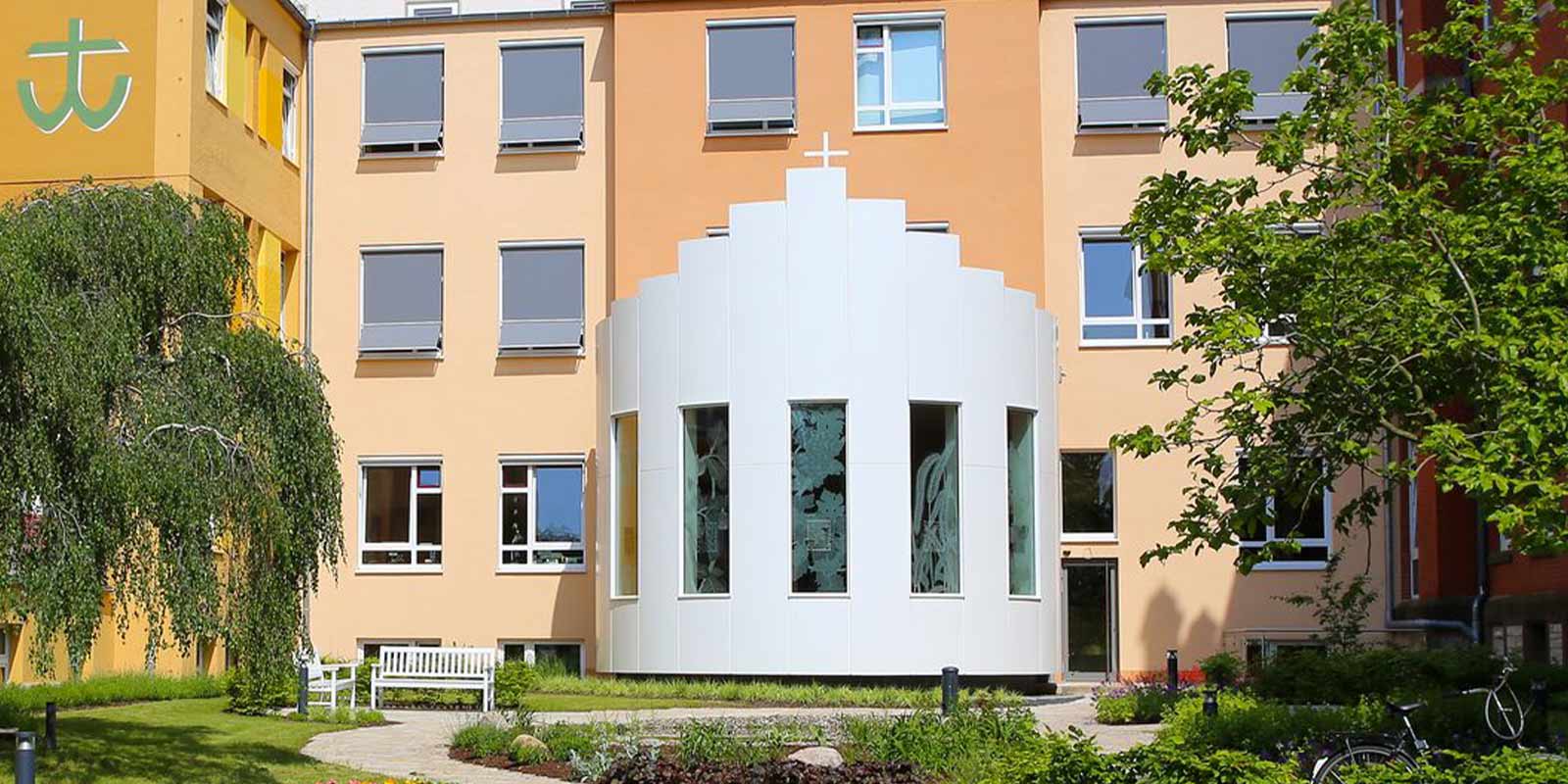 Gelbes Gebäude mit kreuzförmiger Fassade, grüner Hof, Bank und Fahrrad im Vordergrund.