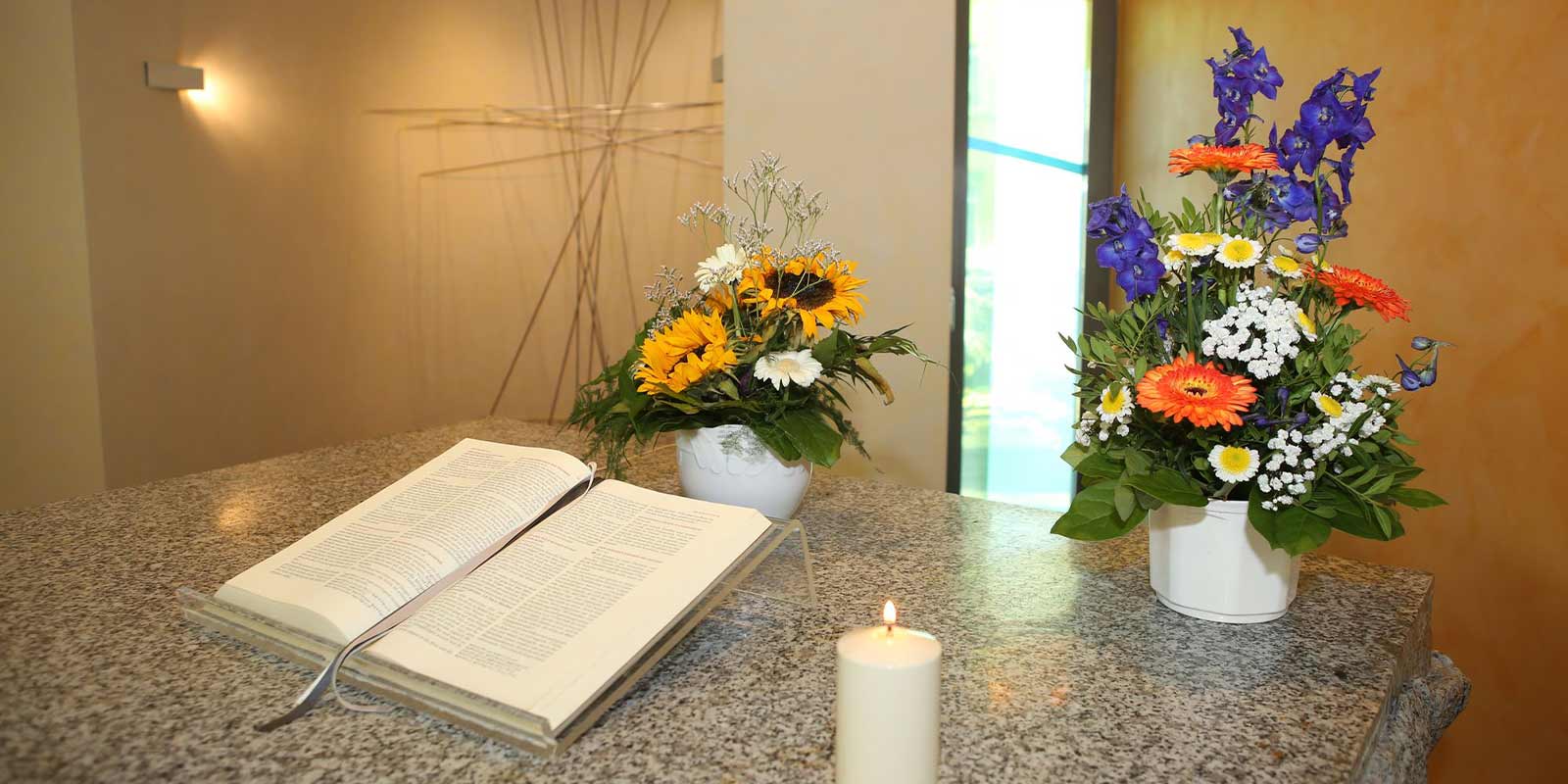 Ein aufgeschlagenes Buch, zwei Blumenarrangements in Vasen und eine brennende Kerze auf einem Tisch.