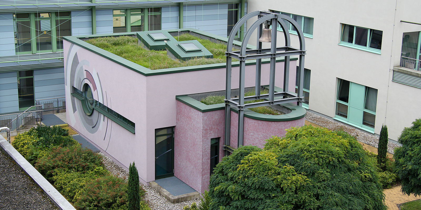 Modernes Gebäude mit Dachbegrünung, Wandkunst und Metallstruktur, umgeben von grüner Vegetation.