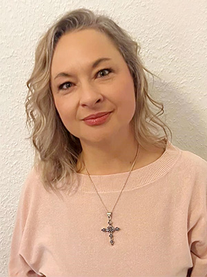 Es zeigt eine Frau mit welligem Haar, rosa Pullover und Halskette mit Kreuzanhänger vor einer weißen Wand.