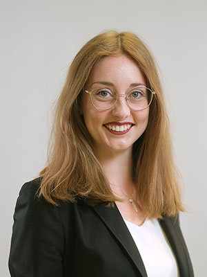 Es zeigt eine Person mit Brille und braunen Haaren in einem schwarzen Blazer, lächelnd vor neutralem Hintergrund.