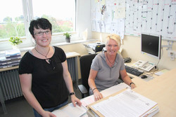Zwei Personen in einem Büro, eine sitzt am Tisch, die andere steht. Computer und Dokumente sind sichtbar.