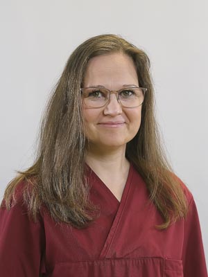 Es ist eine Frau mit Brille, die ein weinrotes Oberteil trägt. Sie lächelt in die Kamera und hat langes Haar.