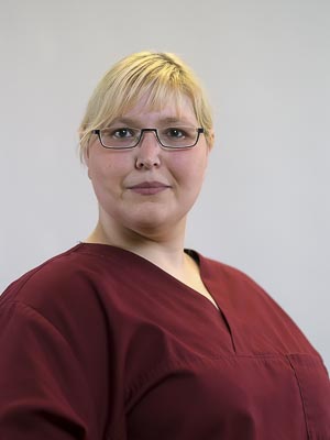 Eine Person mit Brille und blondem Haar trägt rote Arbeitskleidung vor neutralem Hintergrund.