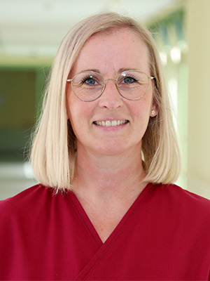 Es zeigt eine lächelnde Frau mit Brille und blondem Haar, die ein rotes Oberteil trägt.