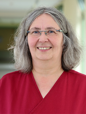 Es zeigt eine lächelnde Frau mit Brille, grauem Haar und roter Oberbekleidung vor verschwommenem Hintergrund.