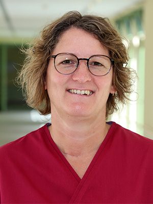 Es zeigt eine lächelnde Person mit Brille und rotem Hemd vor verschwommenem Hintergrund in einem Innenraum.