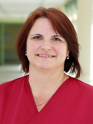 Es ist eine lächelnde Frau mit braunen Haaren und einem roten Oberteil vor einem verschwommenen Hintergrund.