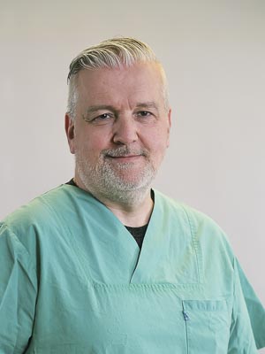 Ein Mann in grüner medizinischer Kleidung vor einem neutralen Hintergrund.