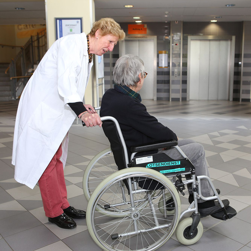 Eine Ärztin schiebt eine ältere Person im Rollstuhl durch ein Gebäude.
