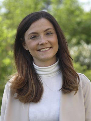 Es zeigt eine lächelnde Frau mit langen Haaren, die einen Rollkragenpullover und eine Jacke draußen trägt.