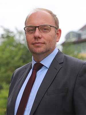 Ein Mann in Anzug und Krawatte steht draußen, trägt eine Brille und blickt in die Kamera.