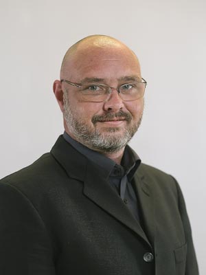 Ein Mann mit Glatze, Bart und Brille trägt ein dunkles Jackett und Hemd, vor neutralem Hintergrund.