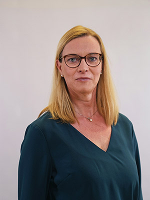 Eine Frau mit blonden Haaren und Brille trägt ein dunkelgrünes Oberteil, vor einem hellen Hintergrund.