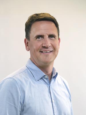 Ein Mann mit kurzem, dunklem Haar trägt ein hellblaues Hemd und lächelt vor einem neutralen Hintergrund.