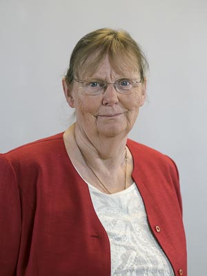 Es ist eine ältere Frau mit Brille in einem roten Blazer vor neutralem Hintergrund.