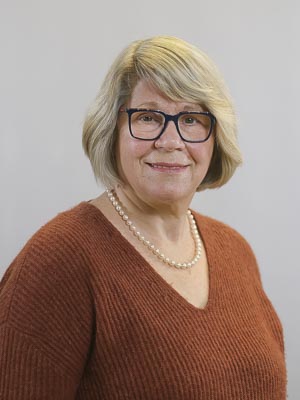 Es ist ein Porträt einer Frau mit Brille, braunem Pullover und Perlenkette vor einem grauen Hintergrund.