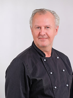 Ein Mann in schwarzer Kochjacke vor einem neutralen Hintergrund.