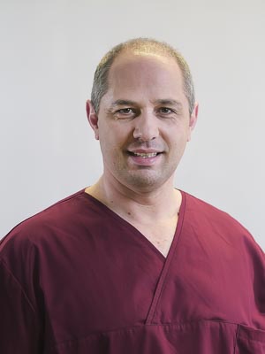 Ein Mann in weinroter medizinischer Kleidung lächelt in die Kamera vor neutralem Hintergrund.