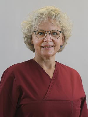 Eine Person mit Brille und lockigem Haar trägt ein weinrotes Oberteil und lächelt in die Kamera.