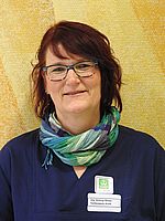 Es ist eine lächelnde Frau mit Brille und einem Schal. Sie trägt ein Namensschild.