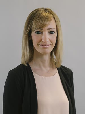 Es zeigt eine lächelnde Frau mit blonden Haaren, die ein rosafarbenes Oberteil und eine schwarze Jacke trägt.