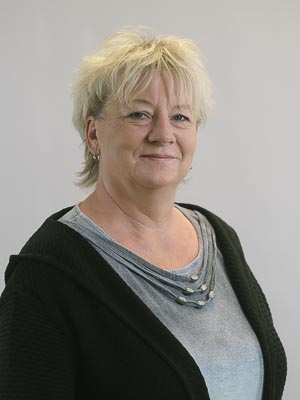 Es ist ein Porträt einer lächelnden Person mittleren Alters mit hellem Haar, die einen schwarzen Pullover trägt.