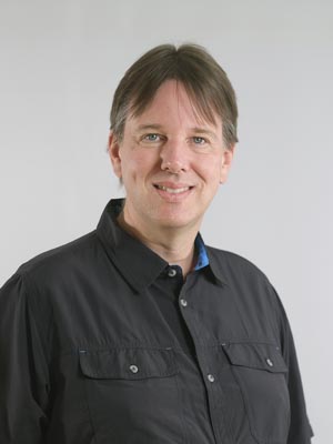 Es ist ein Porträt eines Mannes in einem dunklen Hemd vor einem neutralen Hintergrund.