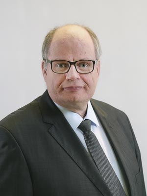 Es ist eine Person in einem Anzug mit Brille vor einem neutralen Hintergrund.