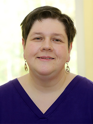 Es ist ein Porträt einer lächelnden Person mit kurzen Haaren und Ohrringen in einem lila Oberteil.