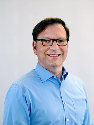 Es ist ein lächelnder Mann mit Brille, der ein blaues Hemd trägt und vor einem hellen Hintergrund steht.