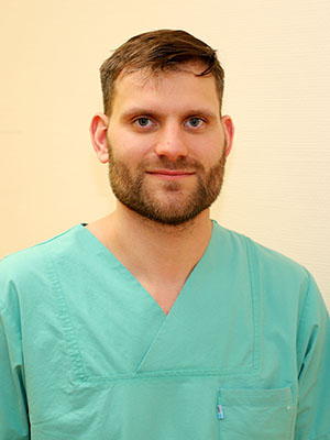 Ein Mann in grüner medizinischer Kleidung steht vor einem neutralen Hintergrund.