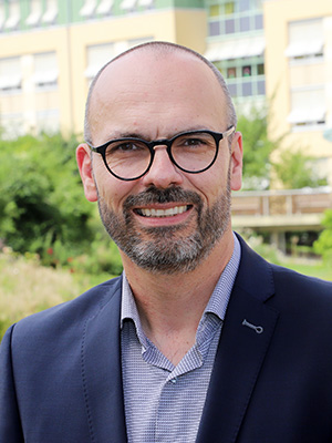 Ein Mann mit Brille und Bart trägt einen Anzug und lächelt in die Kamera. Im Hintergrund ist ein Gebäude.