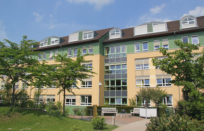 Ein mehrstöckiges Gebäude mit einem gepflegten Garten und Bänken im Vordergrund bei sonnigem Wetter.