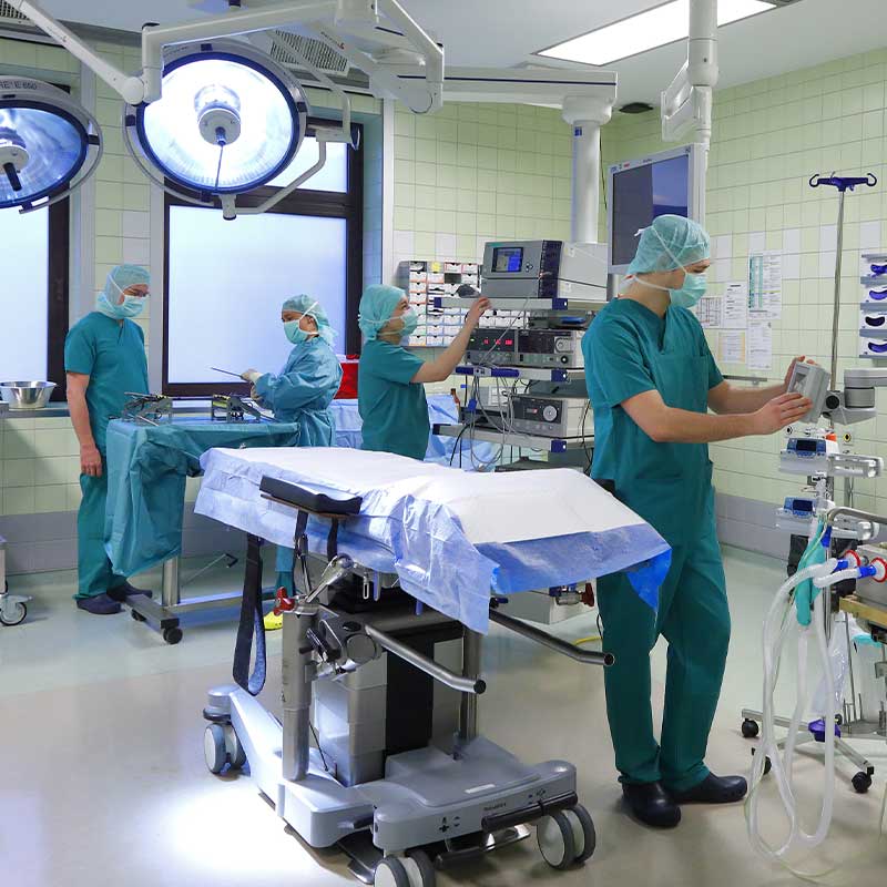 Medizinisches Personal bereitet sich in einem OP-Saal mit Geräten und einem OP-Tisch auf eine Operation vor.